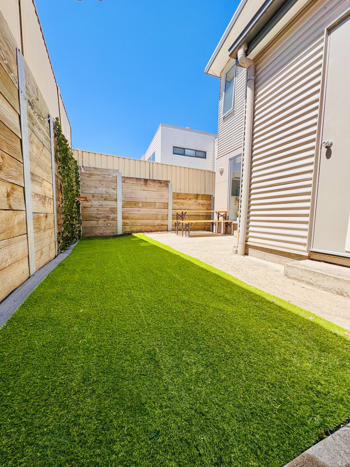 A secure courtyard is shown with artificial grass, bordered by wooden fencing. Sunlight bathes the area, highlighting a small outdoor dining set on a patio. The space offers a welcoming environment for guests and their pets.
