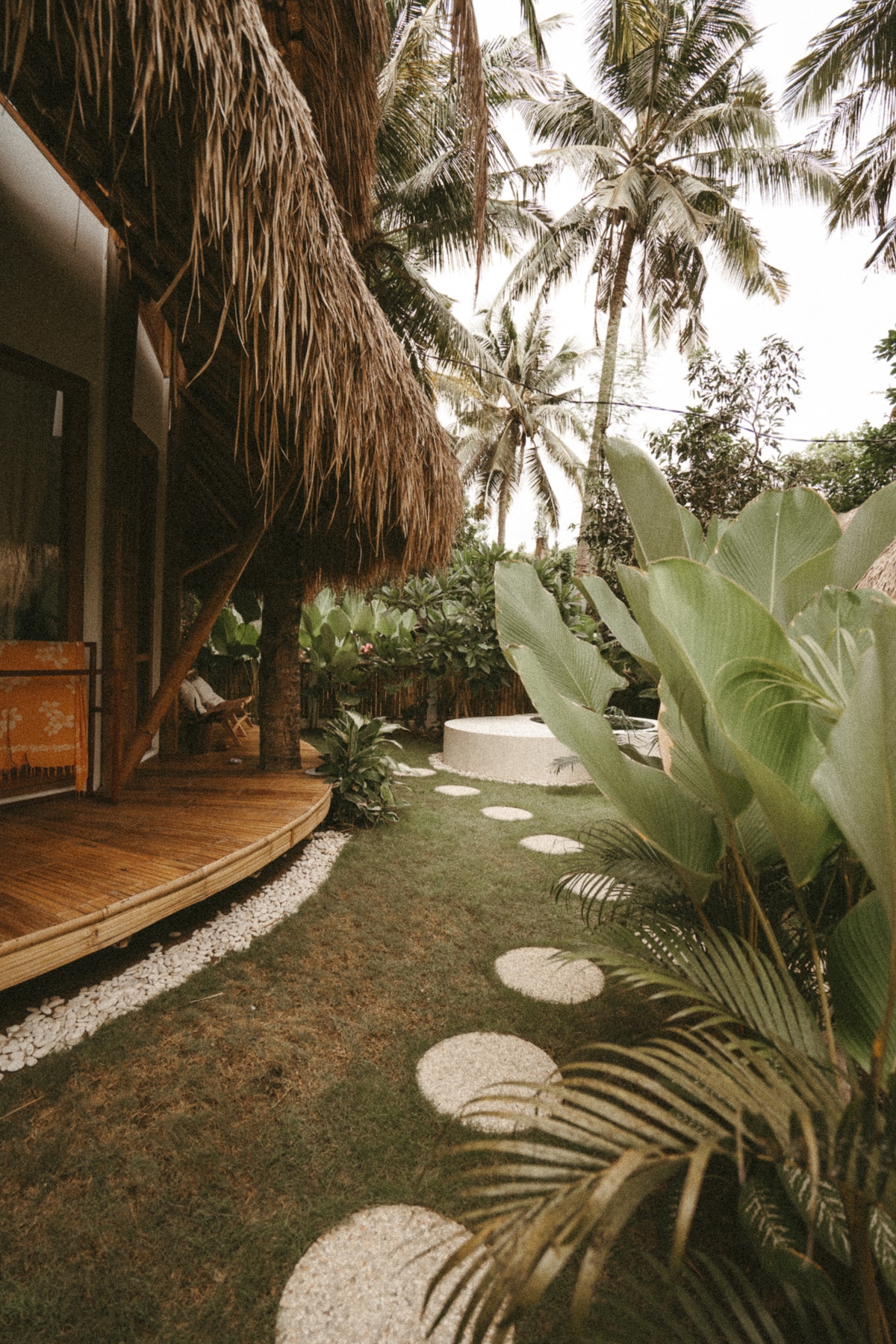 A serene pathway winds through a lush garden, bordered by tropical plants and palm trees. Circular stone steps lead toward a modern white seating area, enhancing the connection with nature. The natural materials of the house's exterior complement the tranquil atmosphere.