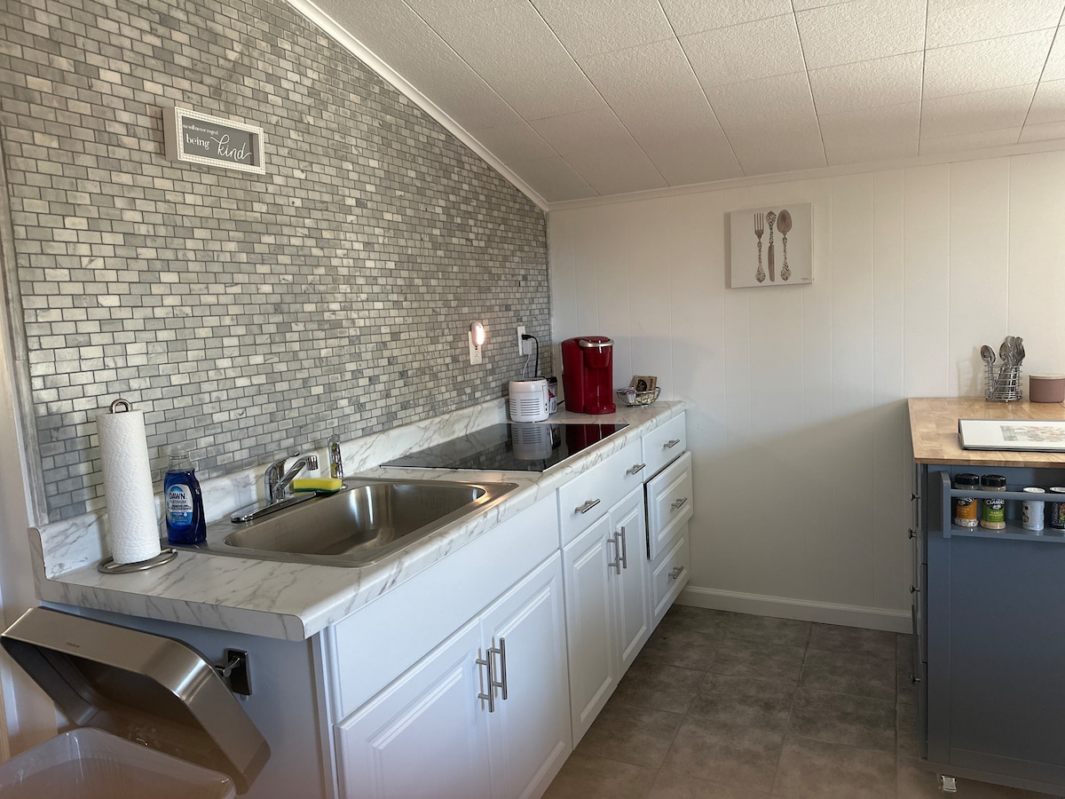 A modern kitchen space features white cabinets and a marble countertop, complemented by a decorative tiled backsplash. Essential appliances, including a coffee maker and stovetop, are visible. A sink and a trash bin are conveniently located, with additional storage cabinets and a small kitchen island in the area.