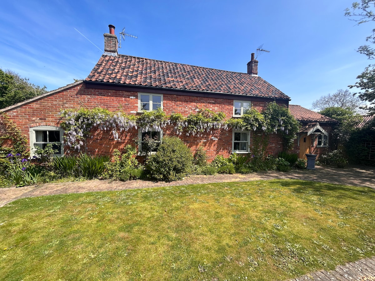 A charming cottage with a red brick exterior is surrounded by lush greenery and flowering vines. A neatly landscaped lawn leads to the entrance, which features a wooden door and a small pathway. The roof is adorned with traditional tiles, complementing the serene environment.