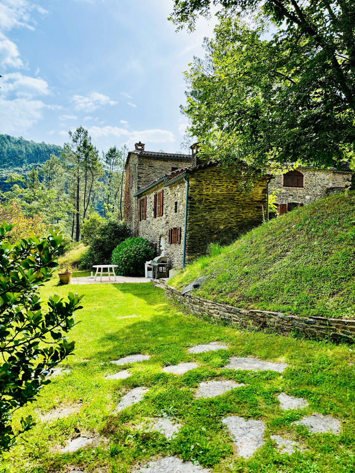 A charming stone cottage is bordered by a lush green lawn, surrounded by trees and hills. A small white table is positioned on the grass, providing a lovely spot for outdoor dining or relaxation. Natural light filters through the foliage, enhancing the serene environment.