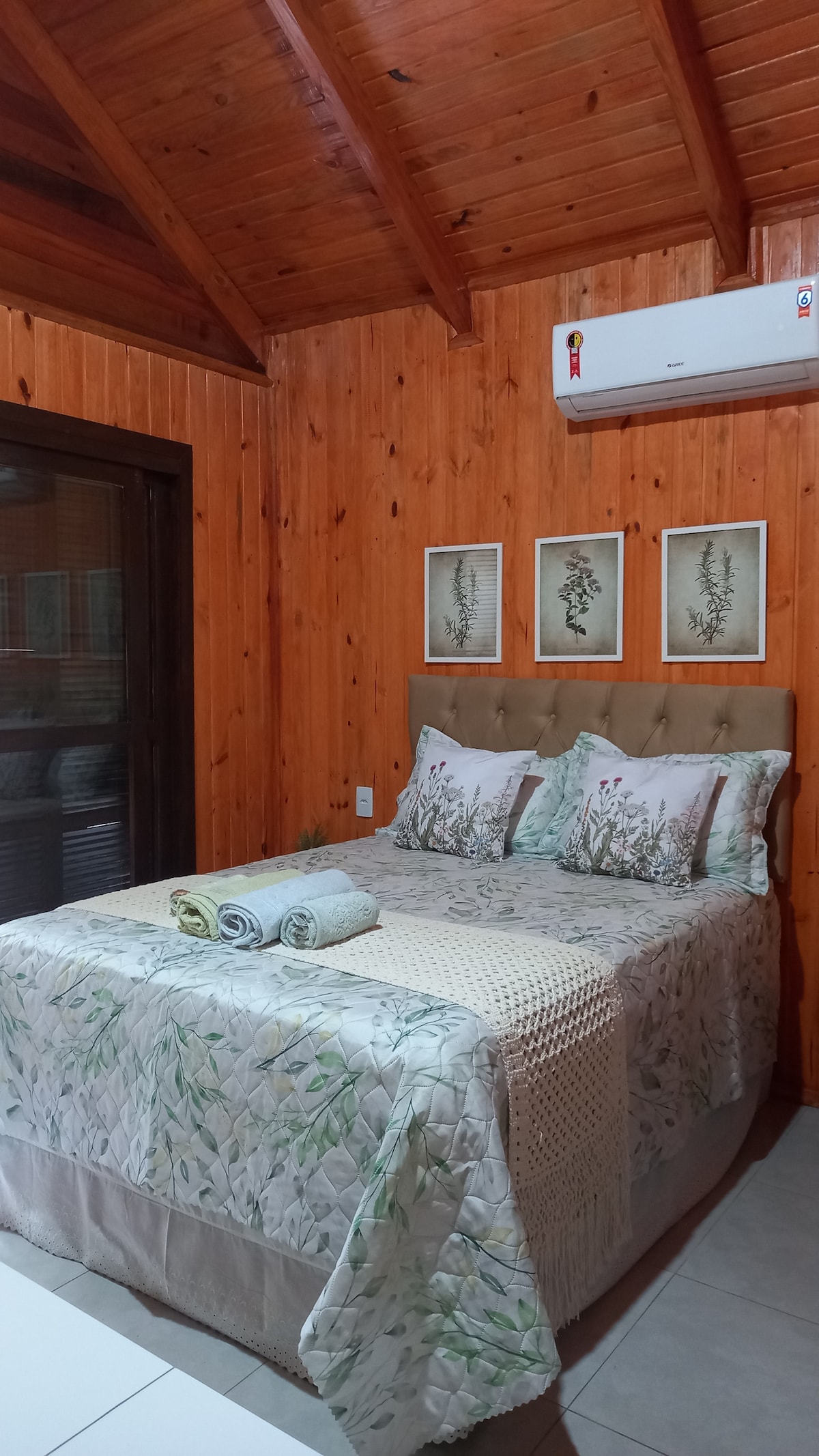 A cozy bedroom features natural wooden walls and a sloped ceiling. A bed with a floral-patterned coverlet is flanked by two framed botanical prints. Soft pillows complement the bedding, while neatly folded towels are placed at the foot of the bed. An air conditioning unit is mounted above.