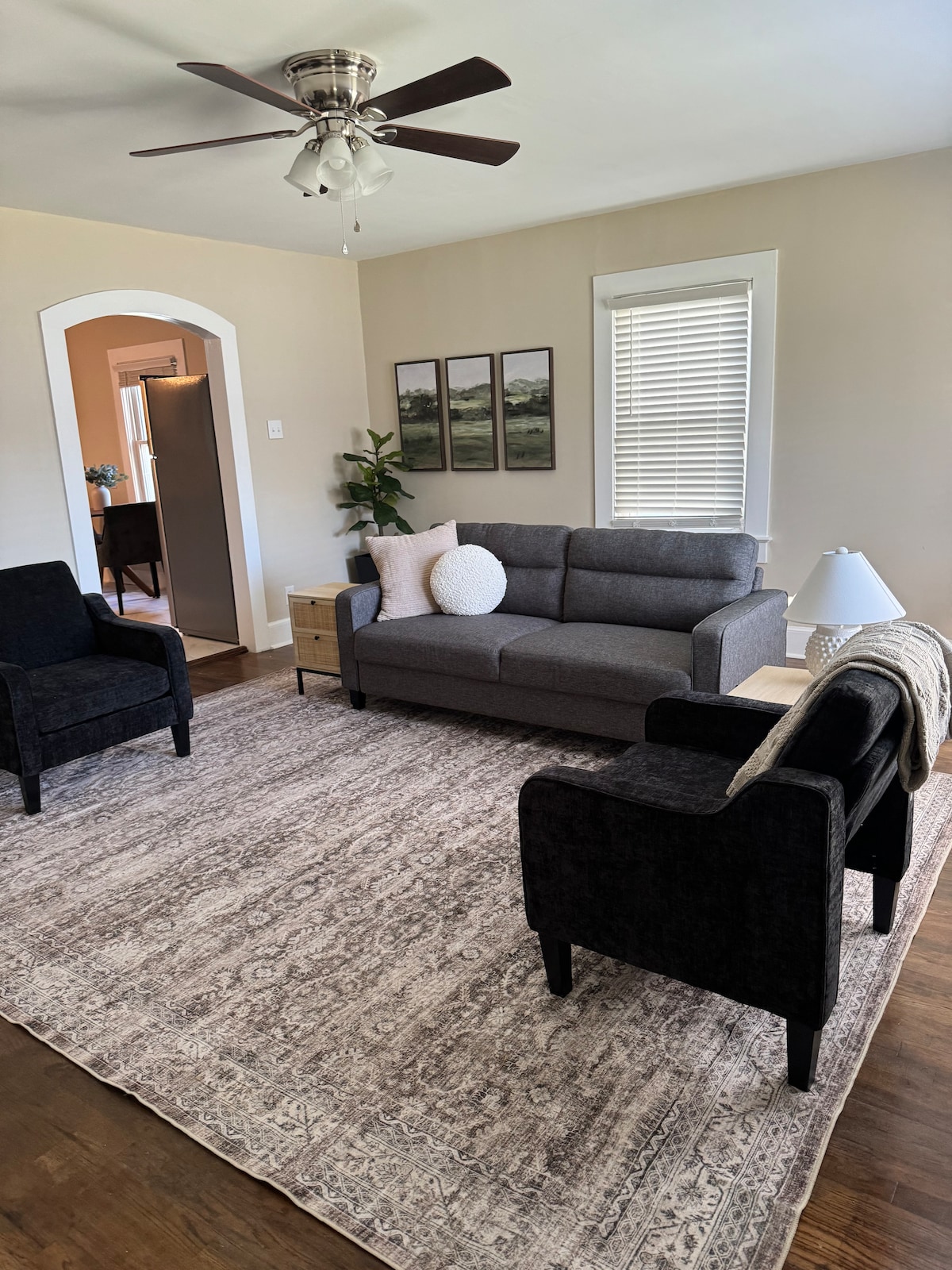 A cozy living area is presented, featuring a gray sofa and two dark chairs arranged on a decorative area rug. Modern art prints hang on the wall, and soft natural light filters through the window, creating a calm environment. A small lamp and pillow add subtle details.