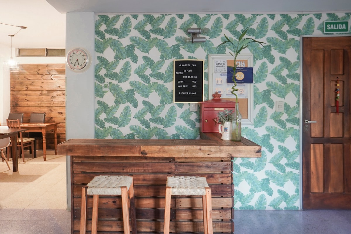 A welcoming entry area features a rustic wooden bar with two simple stools. The wall is adorned with a vibrant leaf-patterned wallpaper. A red mailbox and informational board are noted on the wall, while a clock adds a functional detail to the space.