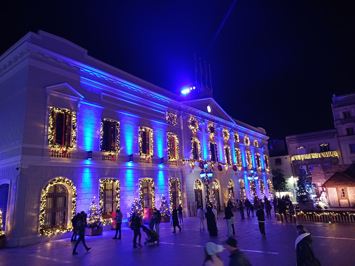 An illuminated building is adorned with blue lights and festive decorations. The facade features multiple arched windows, framed by garlands. People are seen walking in a lively square, with Christmas trees and decorations surrounding the area, creating a cheerful atmosphere.