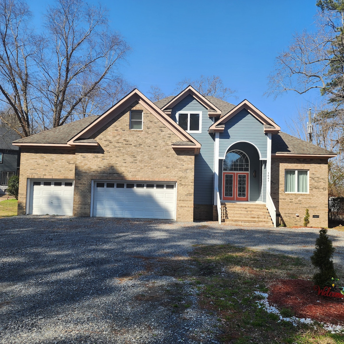 A spacious two-story home features a brick facade and large front steps leading to a central entrance. Flanking the entryway are two oversized garage doors. The surrounding area contains a gravel driveway and trees, creating a welcoming residential setting.