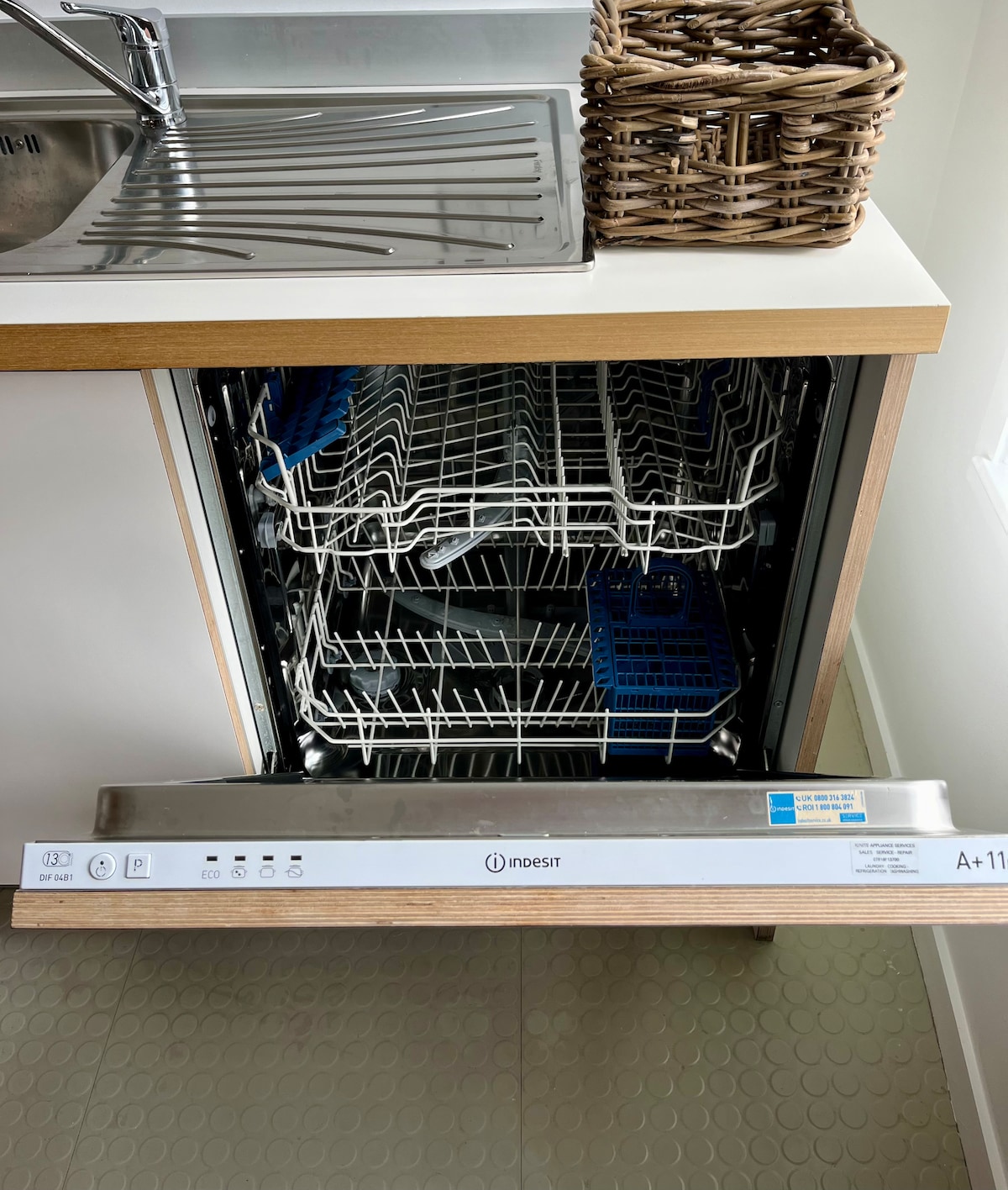 A modern dishwasher is shown with its door open, revealing two spacious racks for dishes. The sleek design features a control panel at the front, and the surrounding cabinetry is finished in light wood tones. A woven basket sits atop the countertop.