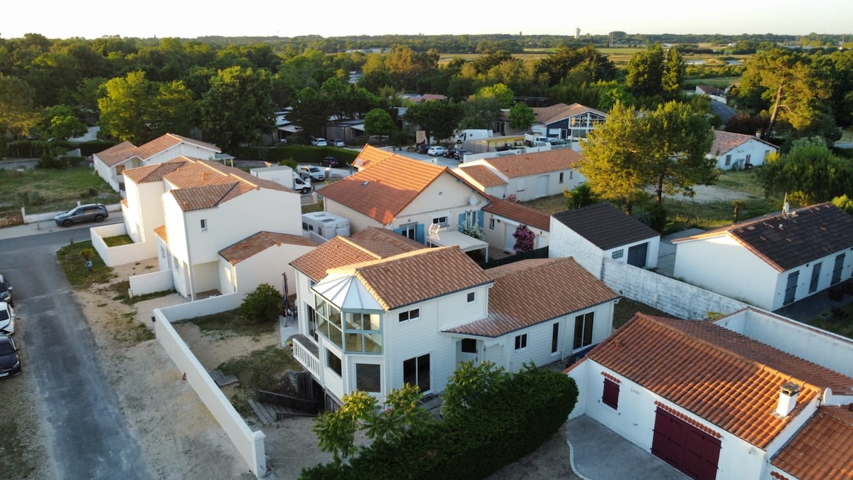 An aerial view captures a collection of homes within a peaceful neighborhood. The distinctive wood-clad house features a glass extension and is surrounded by lush greenery. Adjacent properties exhibit varied roof designs, while a tree-lined landscape provides a serene backdrop.