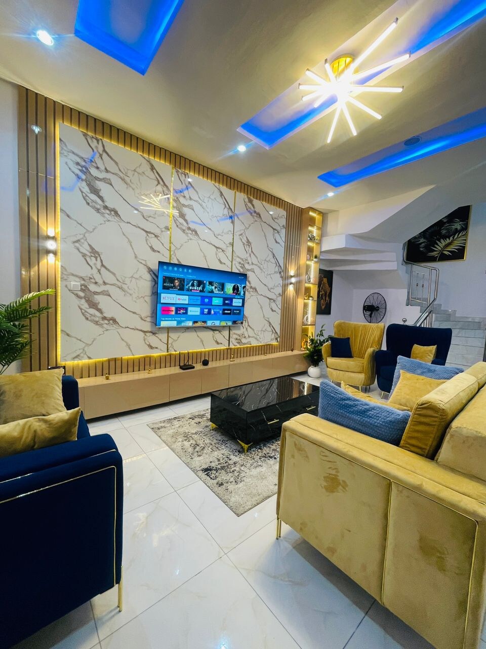 A spacious living area is presented with plush seating in complementary gold and blue tones. An elegant marble feature wall displays a large flat-screen TV, while soft lighting and a decorative ceiling enhance the inviting environment.
