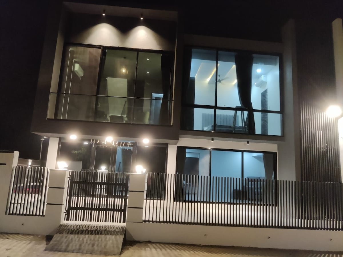A modern villa features large glass windows illuminated by exterior lights, highlighting its contemporary architecture. The façade is complemented by a decorative fence, and the terrace is visible, showcasing a serene outdoor space surrounded by inviting lighting.