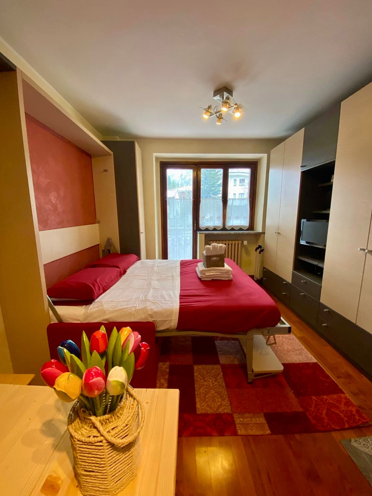 A cozy sleeping area features a spacious bed with a red and white bedding arrangement. A television is situated on a cabinet opposite the bed. Natural light enters through a large window, while colorful flowers in a basket add a touch of vibrancy to the room.