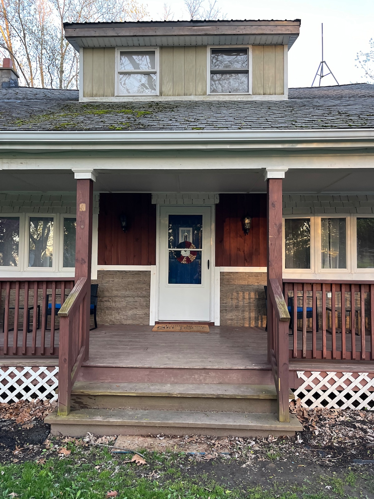 The front of the house features a welcoming porch with wooden railings and steps leading to the entrance. A charming door with decorative elements is framed by light fixtures on either side, complemented by dark wooden accents and a stone foundation. Surrounding greenery adds to the serene setting.
