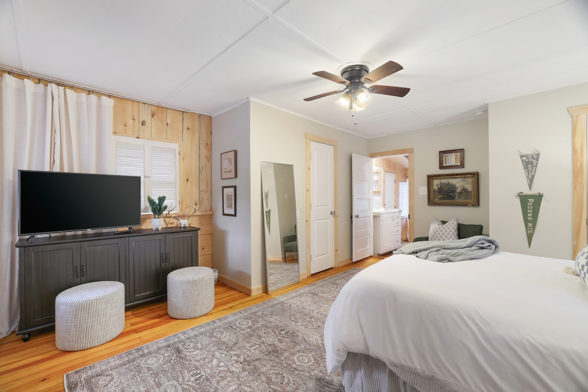 A spacious bedroom features a queen-sized bed with soft linens, complemented by a large mirror and a black media console. Natural light enters through a window adorned with curtains. A playful area rug and two textured stools add comfort to the inviting atmosphere.