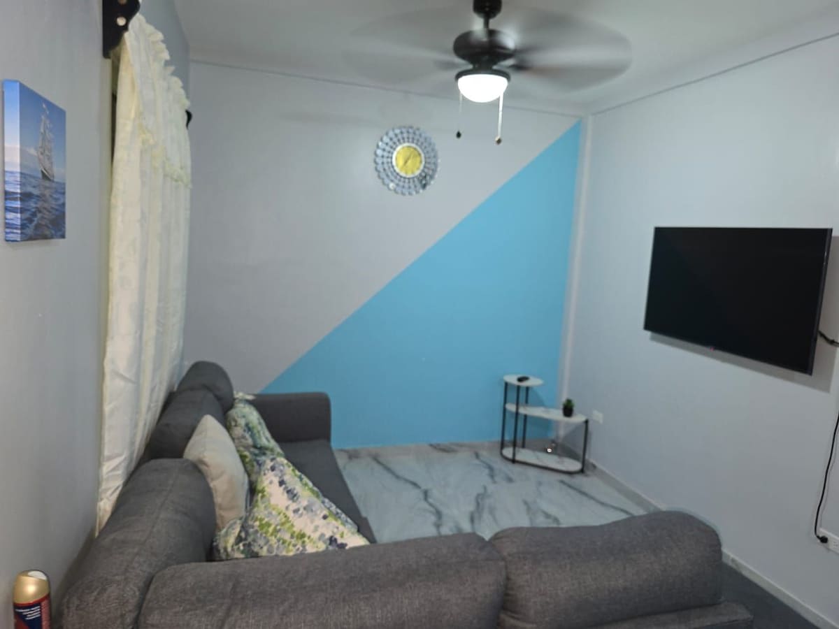 A living area is presented with a gray sectional sofa, accented by decorative pillows. A mounted flat-screen television is visible on one wall. A ceiling fan circulates air while a small side table sits nearby, completing the cozy space decorated with soft blue and white tones.