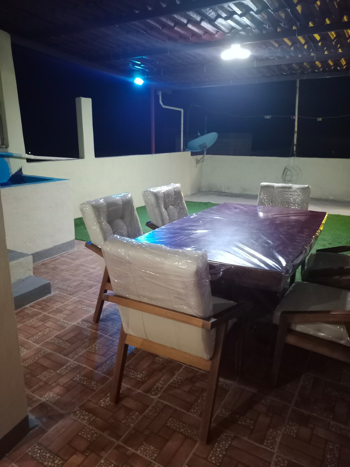 An outdoor dining area is presented, featuring a large table surrounded by several cushioned chairs. The space is illuminated by overhead lighting, with green grass flooring and a partial view of a nearby satellite dish.