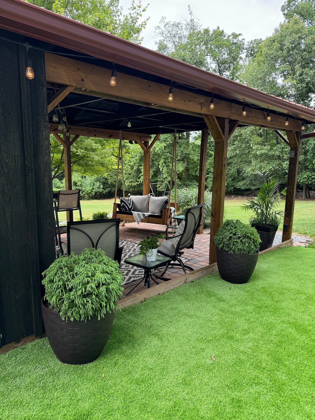 A spacious outdoor pavilion features comfortable seating arrangements, including a cozy farmhouse swing. Elegant planters filled with lush greenery enhance the inviting atmosphere. Soft string lights are suspended overhead, creating a welcoming space for relaxation and gatherings.