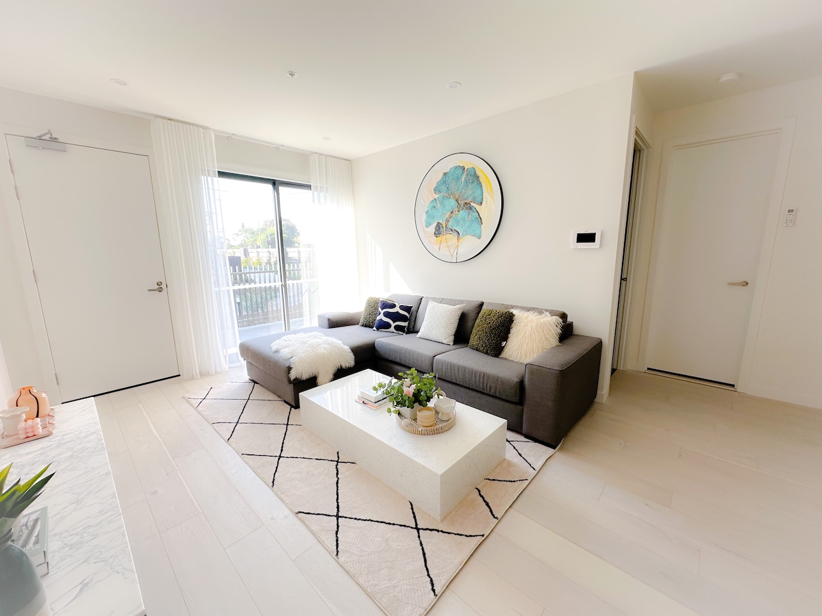The living area features a spacious sectional sofa adorned with decorative cushions. A modern coffee table sits atop a soft area rug. Large glass doors allow natural light to flow in, leading to a balcony with views of the surroundings.