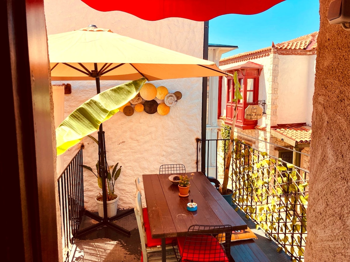 A charming terrace is showcased, featuring a wooden dining table surrounded by colorful chairs. An oversized umbrella provides shade, and potted plants add greenery. The background reveals historic stone buildings and vibrant window frames, lending a lively atmosphere to the outdoor space.