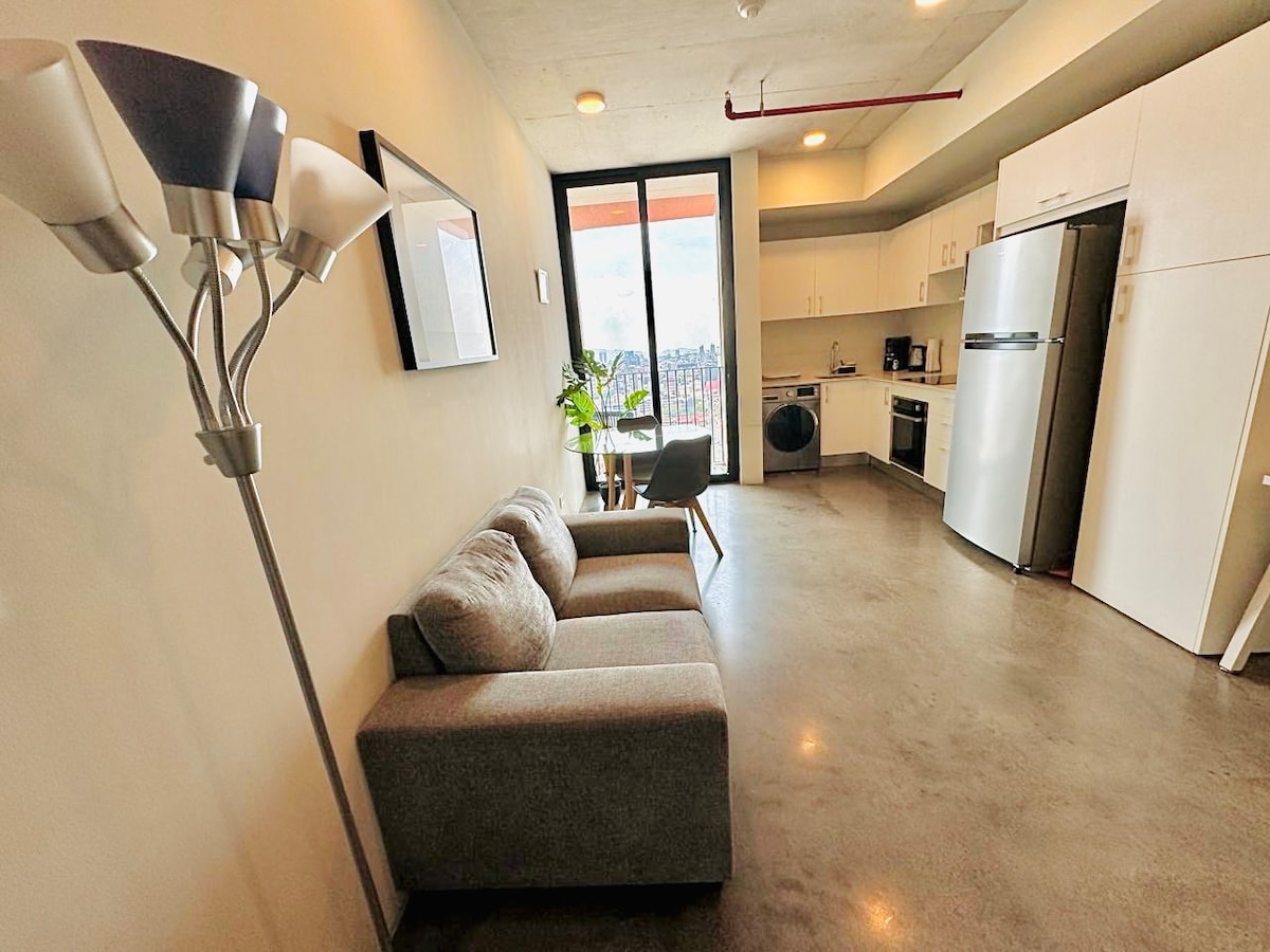A modern studio apartment is featured, showcasing a comfortable gray sofa paired with a floor lamp. The kitchen is equipped with white cabinetry and stainless-steel appliances. Large windows provide natural light and views of the surrounding area, creating an open and airy feel.