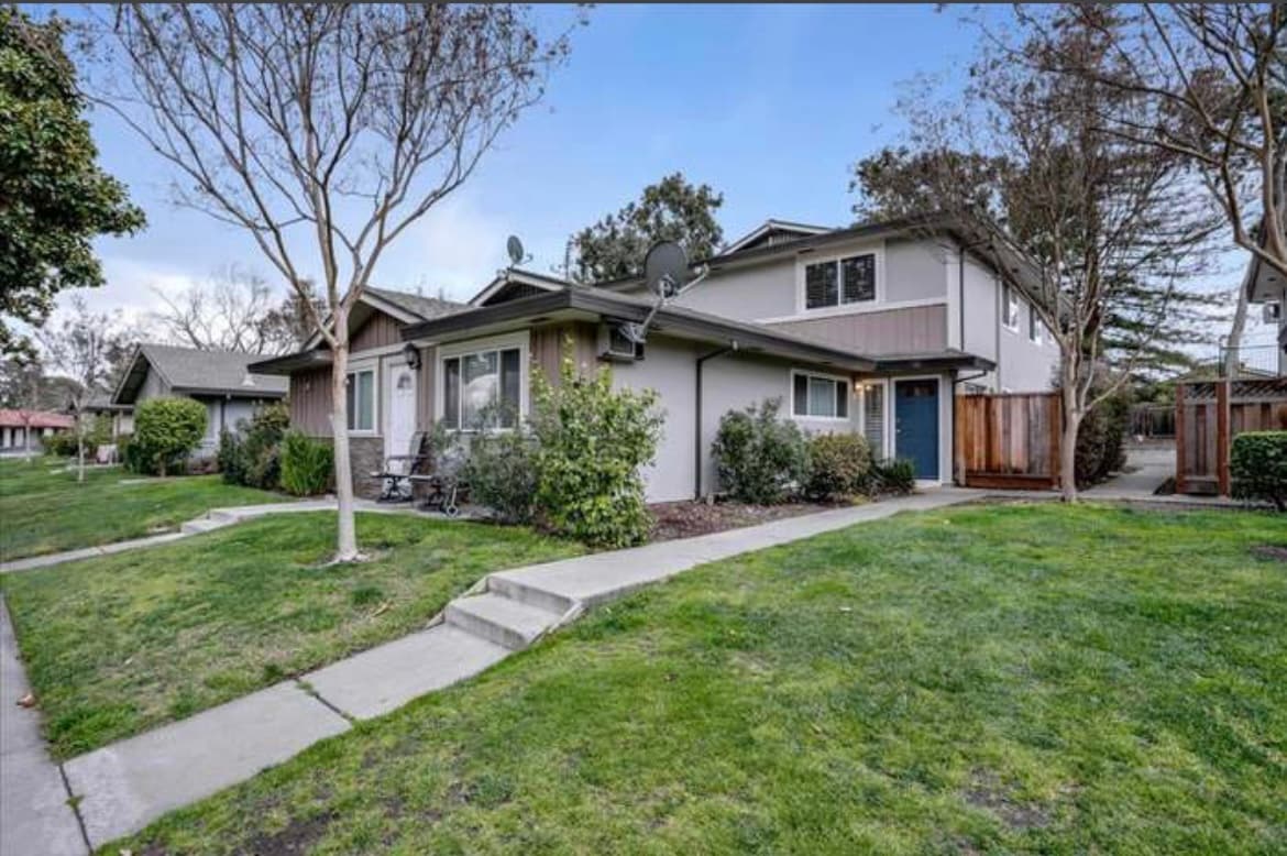 The exterior of a two-story townhouse-style condo is depicted, featuring a combination of light and dark siding. Well-maintained lawns with small trees and shrubs surround the building. A pathway leads to the entrance, which is framed by greenery and modern landscaping.