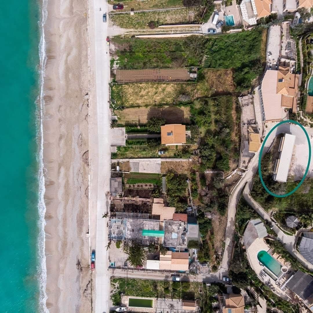 An aerial view captures the coastline adjacent to the accommodation. Clear turquoise waters meet sandy beaches, lined with residential properties and lush greenery. The layout of the local area is visible, including nearby structures and pathways leading to the beach.