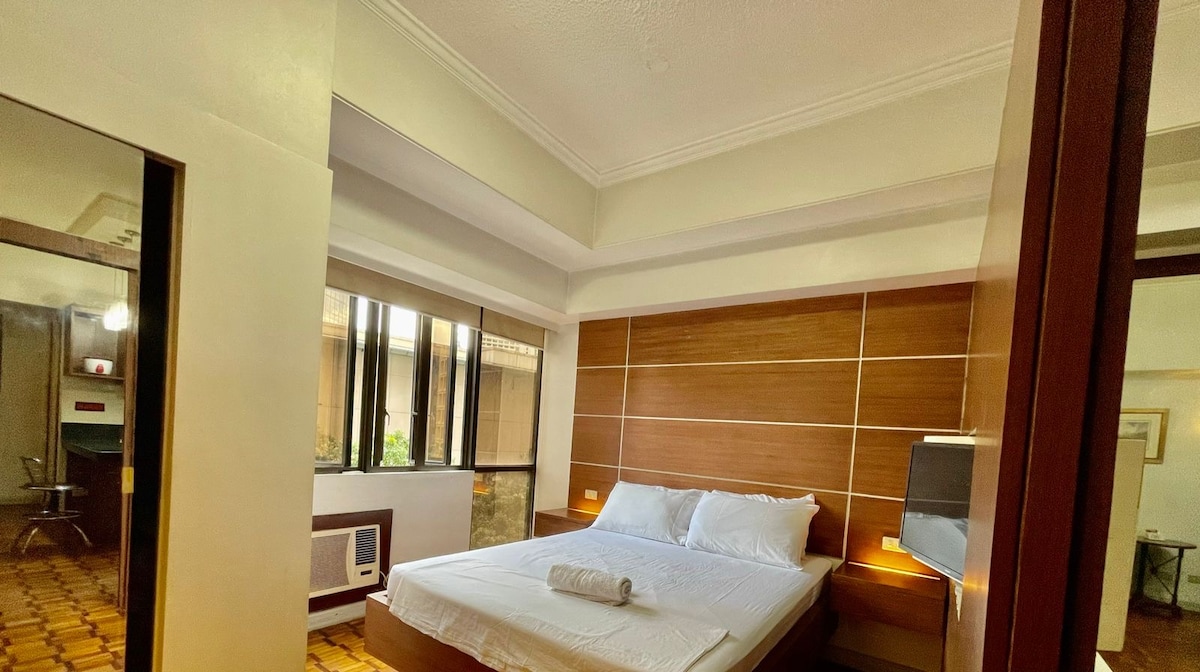 A well-furnished room is highlighted by a spacious bed with crisp white linens. Natural light spills in through large windows, while wood paneling adds a touch of warmth. A sleek work desk can be seen in the background, enhancing the room's functionality.