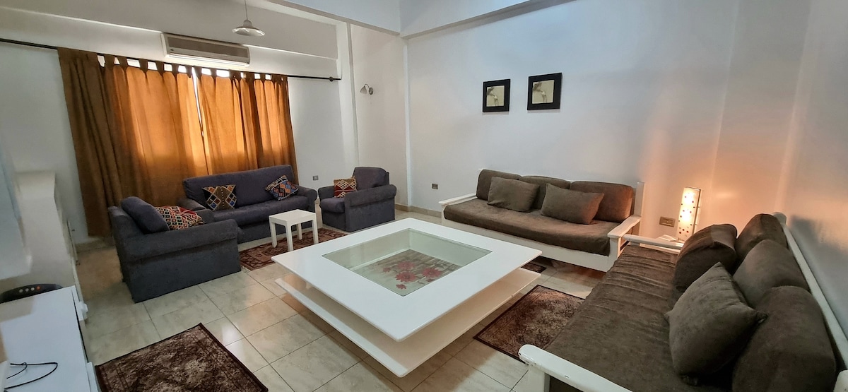 The living area features a light-colored, rectangular coffee table at the center, surrounded by plush, dark sofas. Natural light filters in through large windows covered with warm-toned curtains, while framed wall art adds a touch of style to the space.