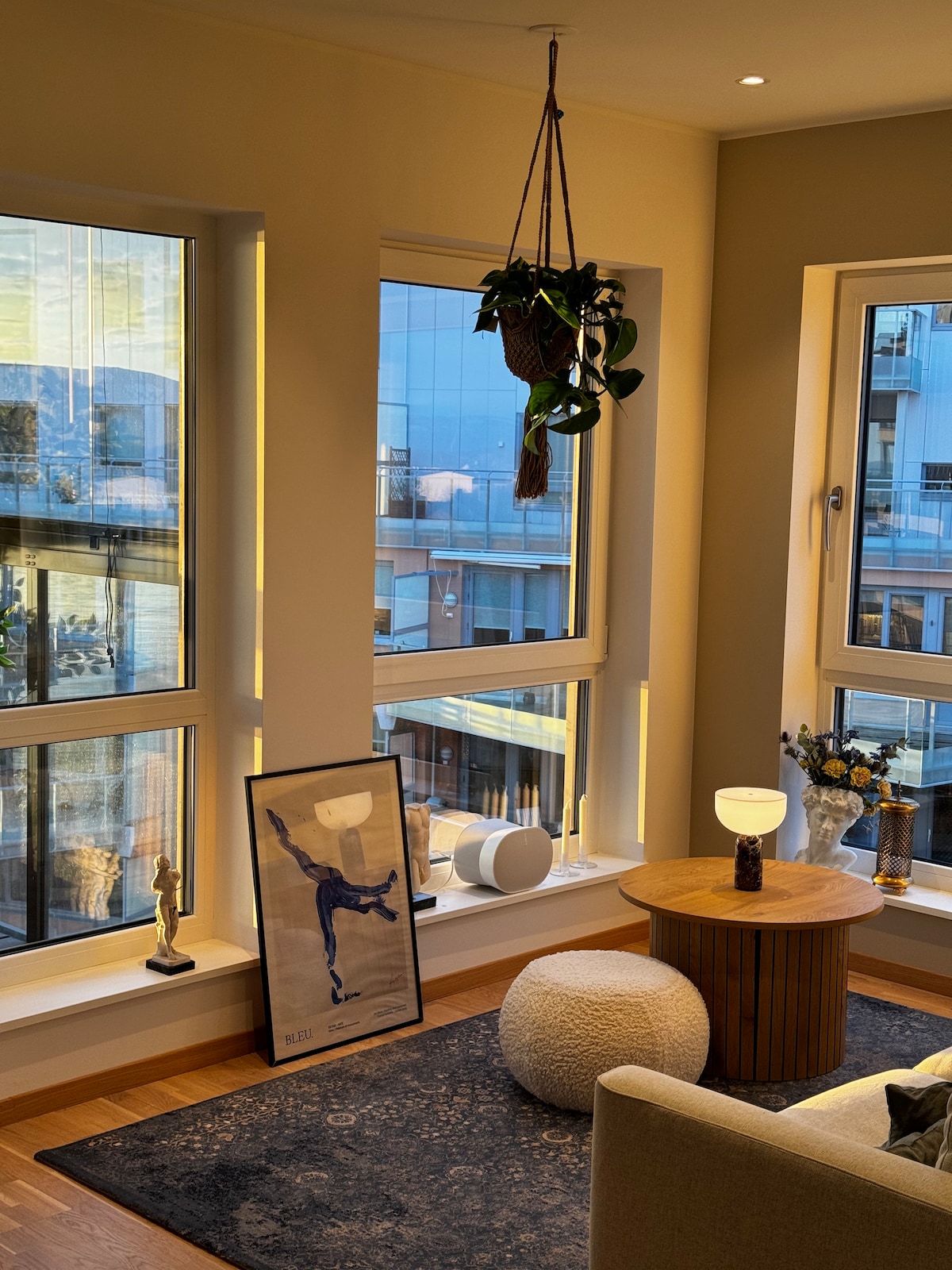 Bright living area features large windows that provide abundant natural light and views of the surroundings. A potted plant hangs from the ceiling, while a wooden table and decorative items, including a framed artwork and a soft pouf, are positioned on a textured rug.