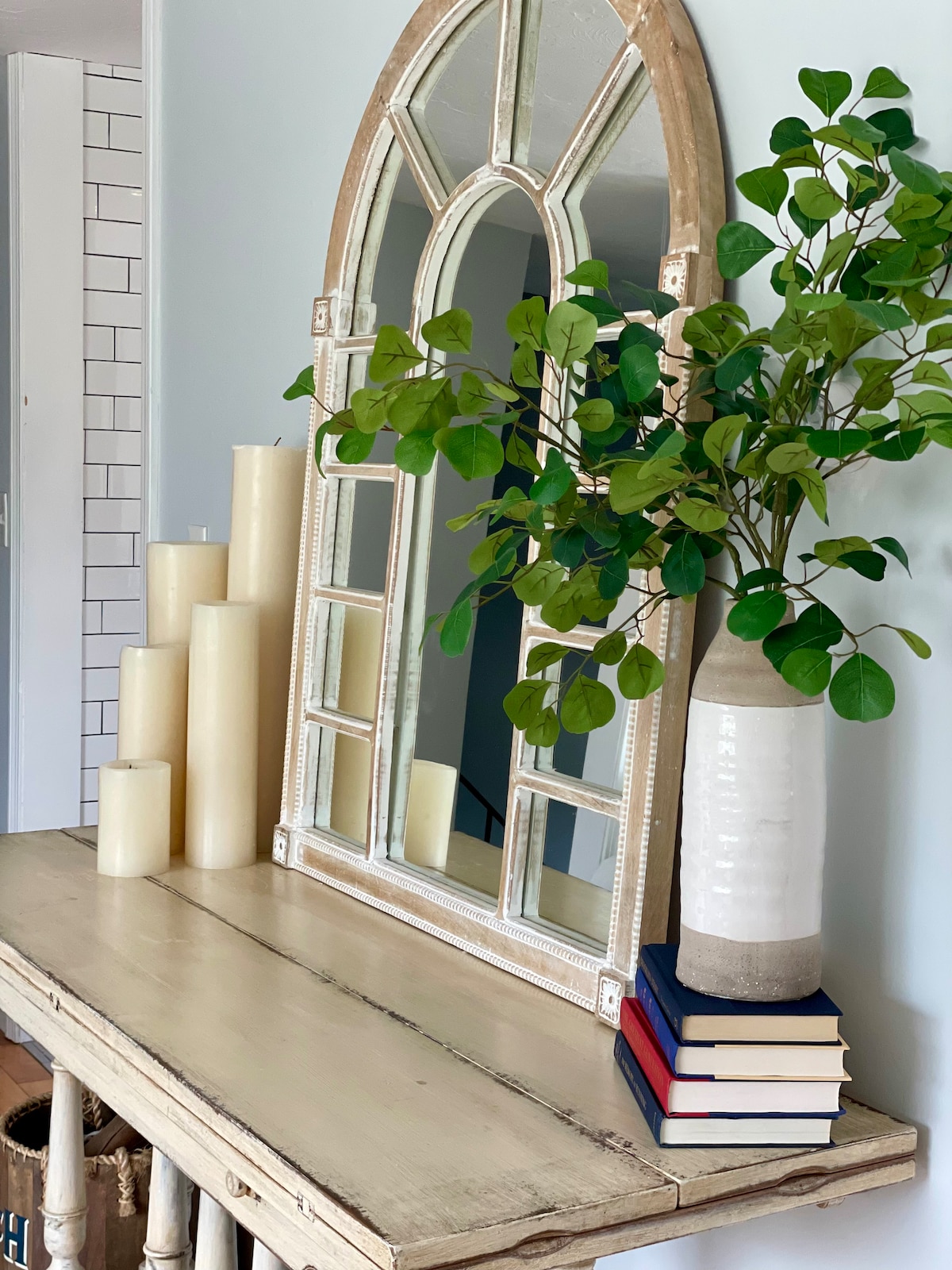 A wooden console table displays a large arched mirror, paired with four cream-colored candles of varying heights. A vase holding leafy greenery rests next to a stack of three books, creating an inviting focal point in the space.
