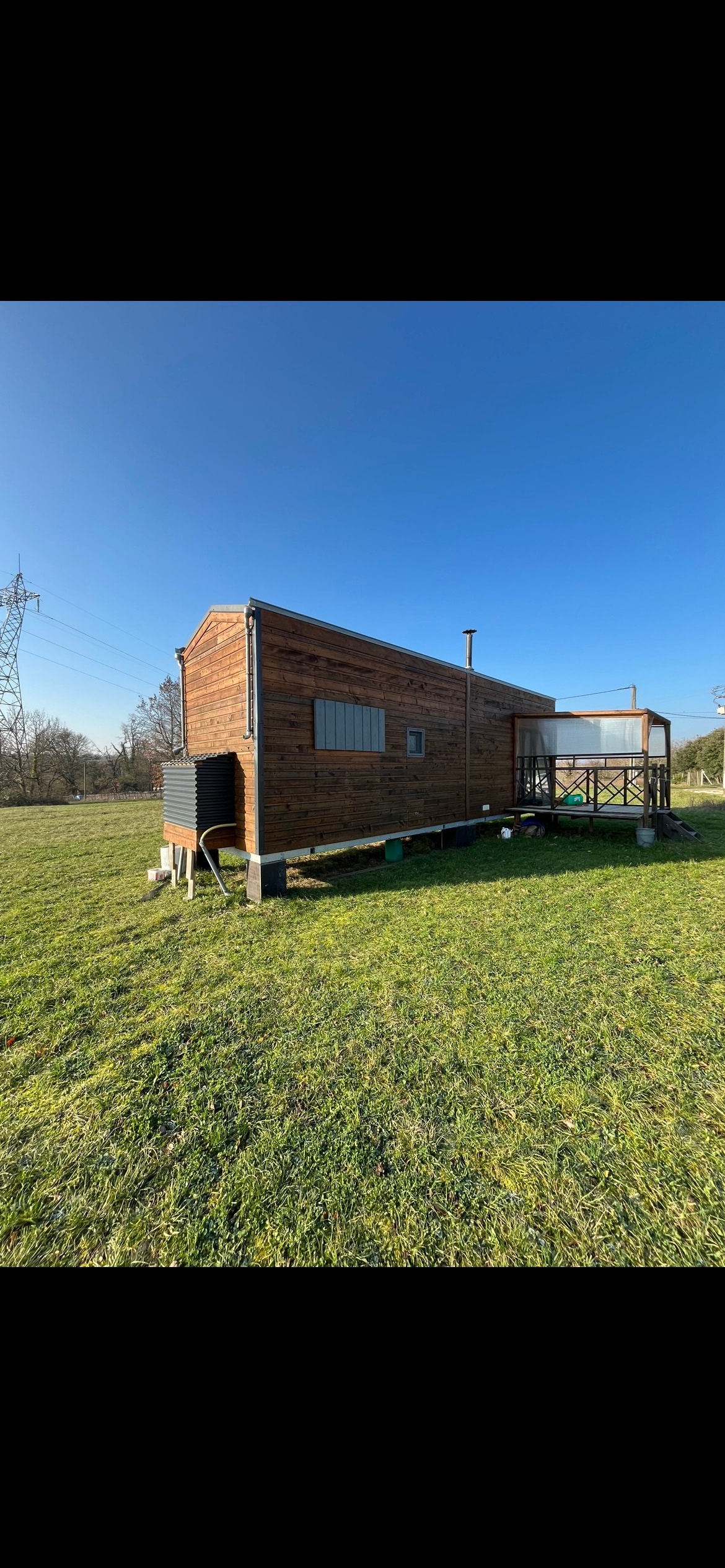 A tiny house made of wood is set within a grassy area under a clear blue sky. The structure features large windows and a deck. The surrounding landscape is open, providing a sense of spaciousness.