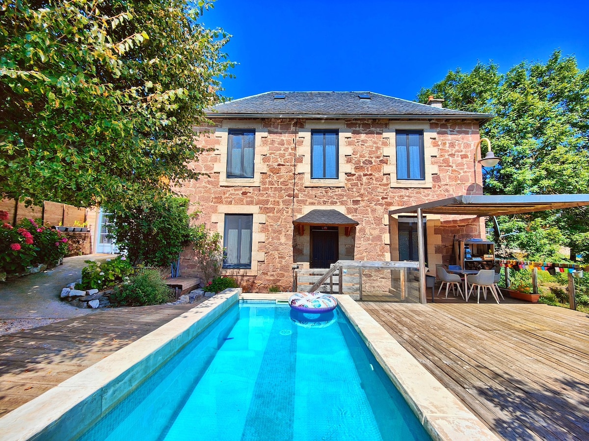 A stone house is depicted with two floors, framed by lush greenery and colorful flowers. A narrow swimming pool is visible in the foreground, alongside a wooden deck with shaded seating. Large windows allow natural light to fill the interior spaces.