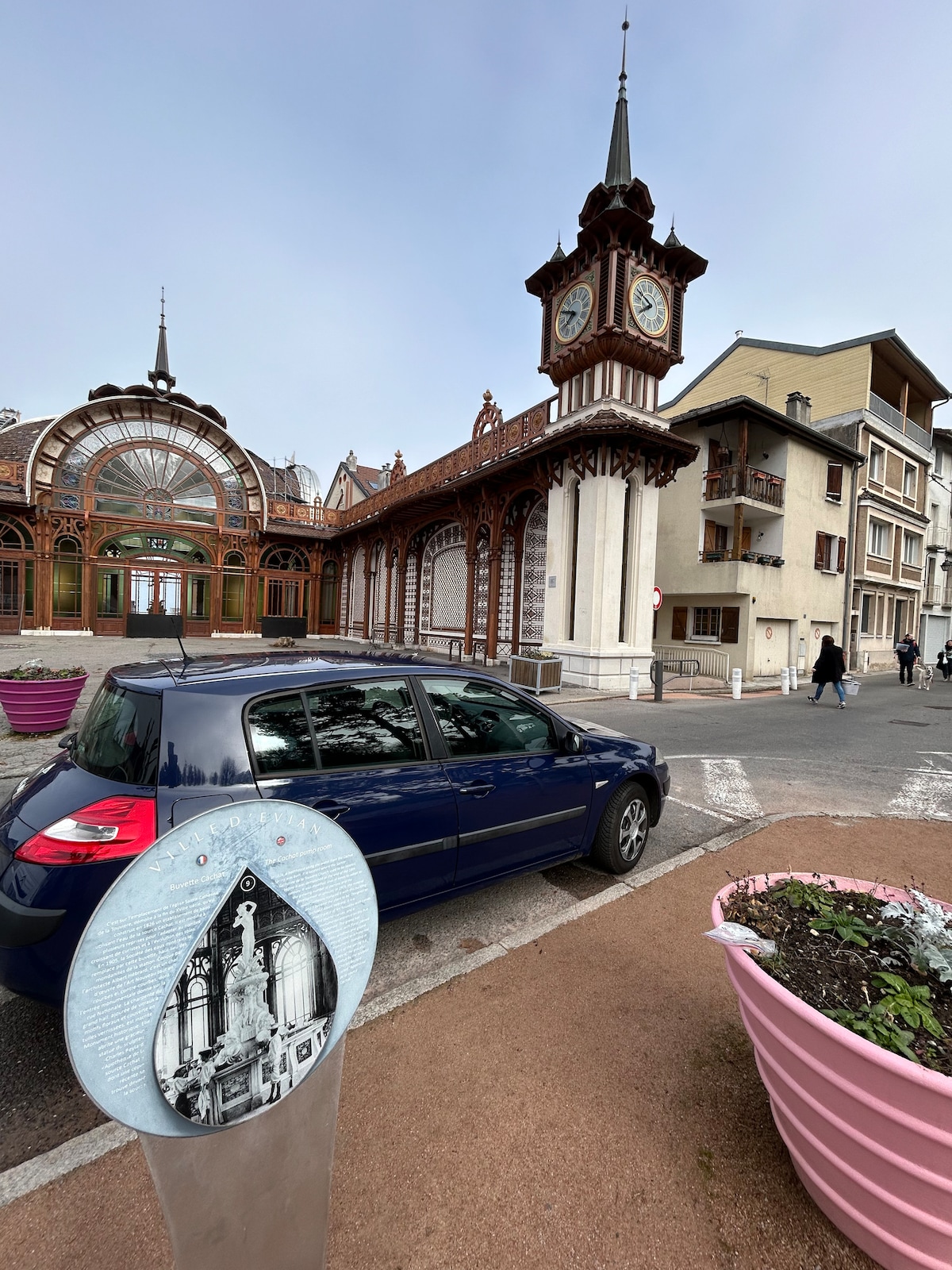 An elegant clock tower is observed in the background, showcasing intricate architectural details. A vintage-style decorative sign is positioned in the foreground, while a parked blue car is visible nearby. The scene captures the charm of historical buildings alongside urban elements.