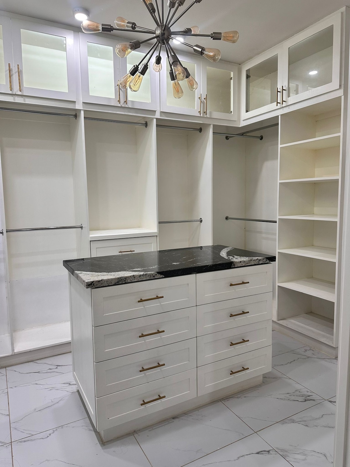 A spacious walk-in closet is equipped with several shelves and hanging areas. A central island features multiple drawers with sleek gold handles and a black countertop. Bright ceiling lights illuminate the organized space, enhancing functionality.