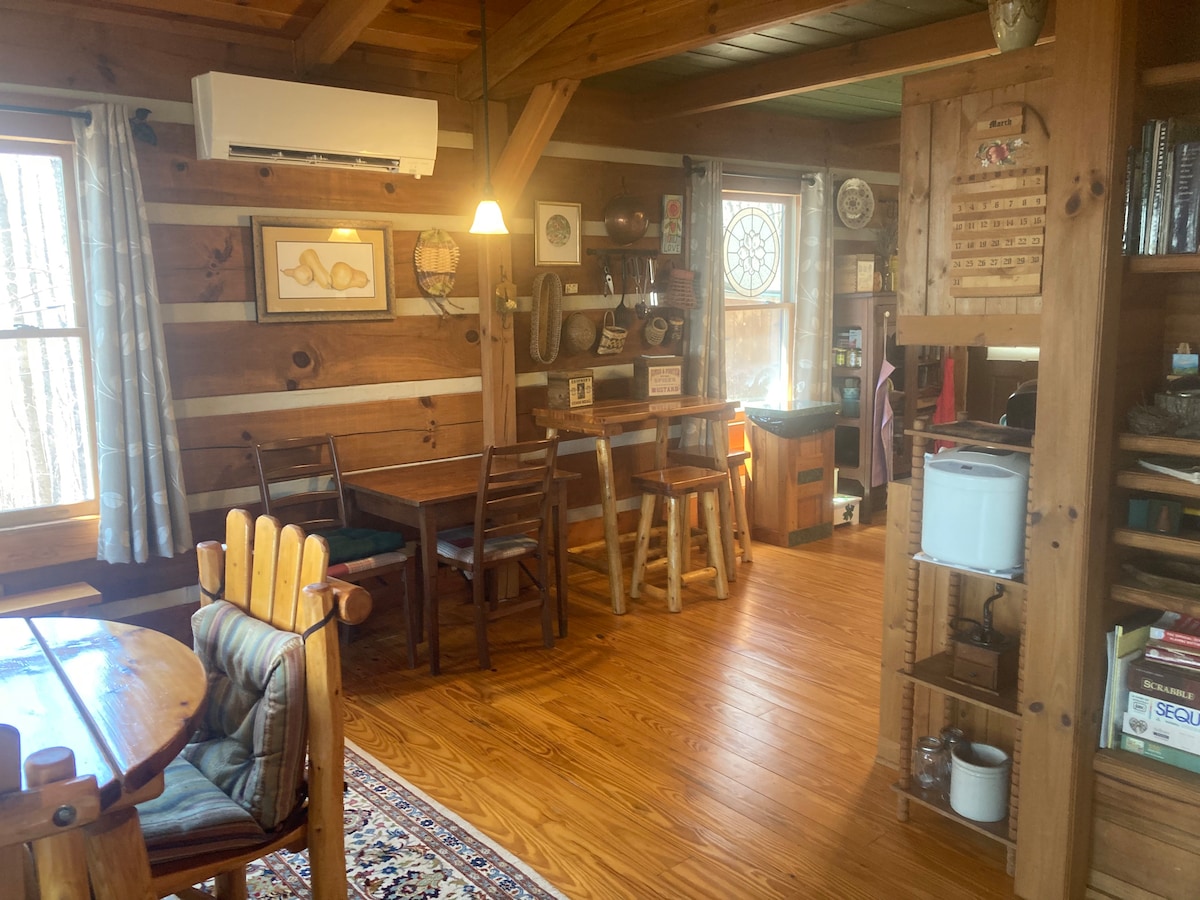 A warm, inviting interior is captured, showcasing wooden beams and paneling. A dining table with four chairs is visible alongside a console table. A well-equipped kitchen can be seen in the background, complemented by a bright window that allows natural light to fill the space.