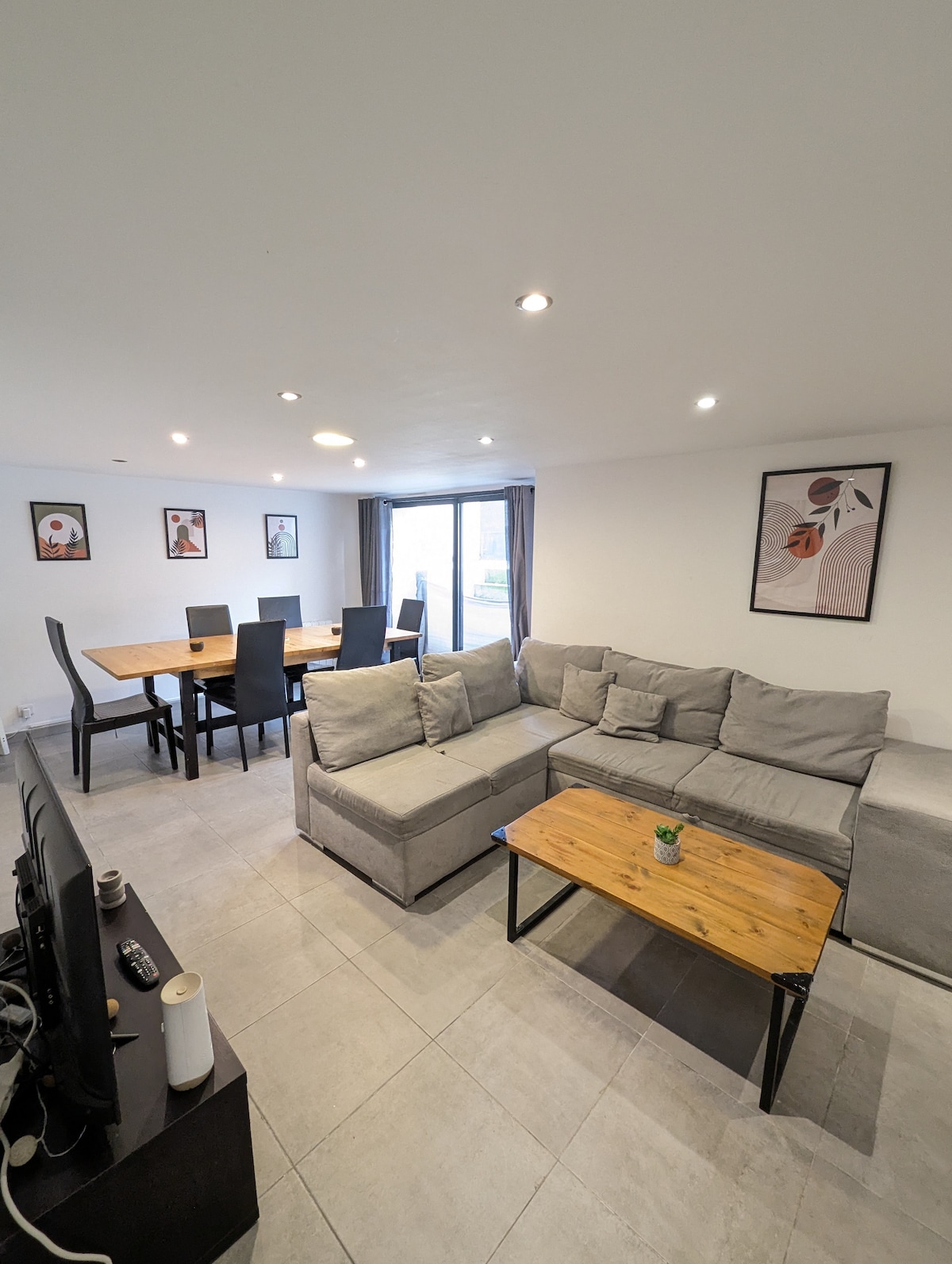 A living area features a spacious L-shaped gray sofa, accompanied by a wooden coffee table. A dining table with six chairs is visible. Light enters from a large window, partially covered by dark curtains. Framed art pieces adorn the walls.
