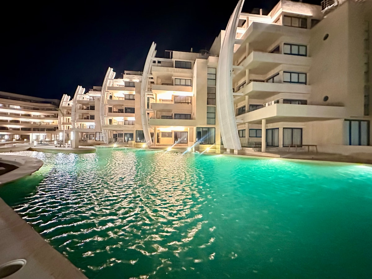 The illuminated pool reflects the soft glow of surrounding lights, creating a tranquil atmosphere. The curved architectural features of the buildings resemble sails, enhancing the unique design. Spacious terraces are visible, providing access to the pool area, which is surrounded by a smooth deck.