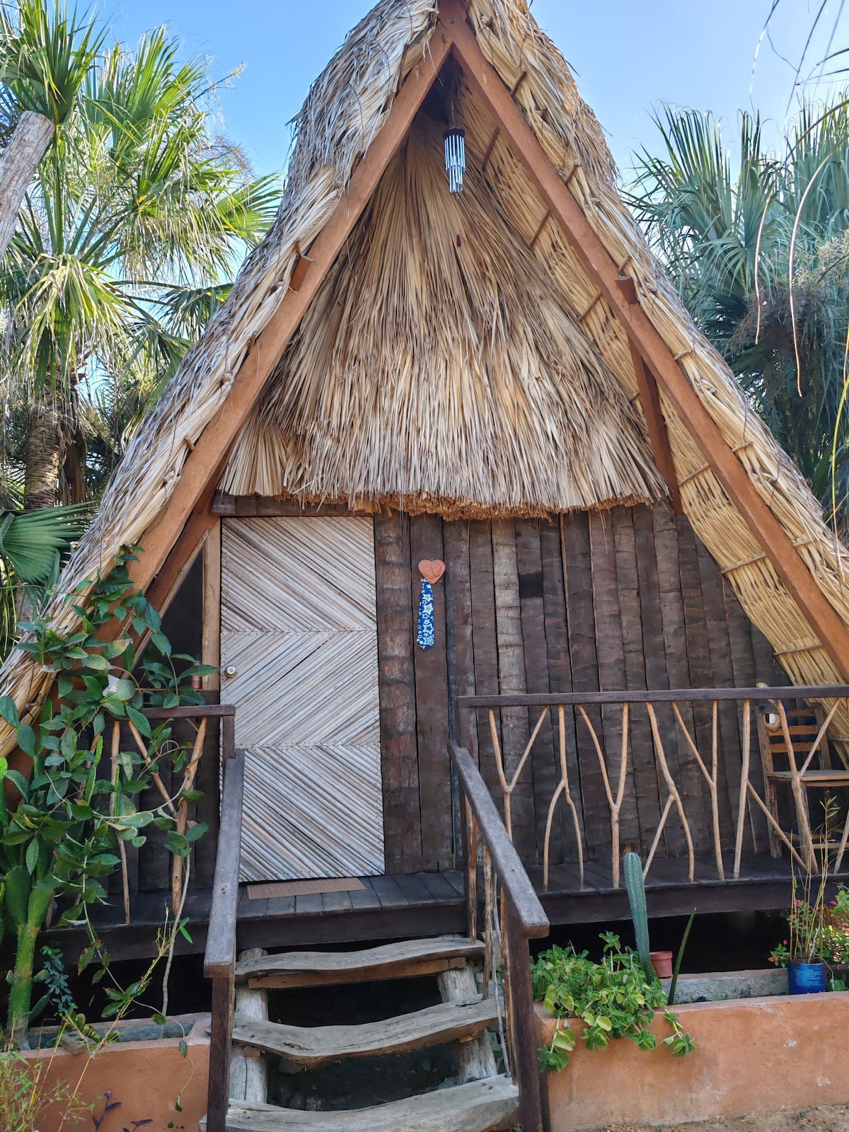A charming triangular-shaped structure features a thatched roof and wooden accents. A decorative door with a textured design is visible, complemented by a small porch with wooden railing and steps leading to the entrance. Lush greenery surrounds the exterior.