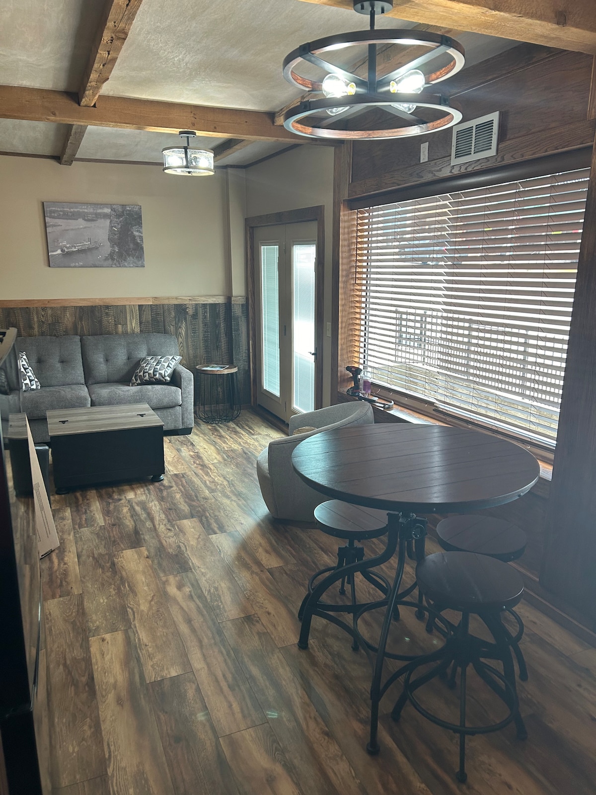 A cozy living area is featured with a modern gray sofa and matching armchair, complementing a round dining table. Natural light filters through window blinds, illuminating the wooden floors and rustic wooden accents throughout the space.