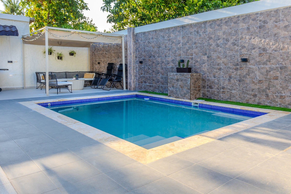 A private pool is surrounded by a tiled patio and stone wall, featuring steps leading into clear water. Relaxation spaces are offered with lounge chairs and a shaded area covered by a canopy. Ambient lighting adds a serene touch to the outdoor setting.