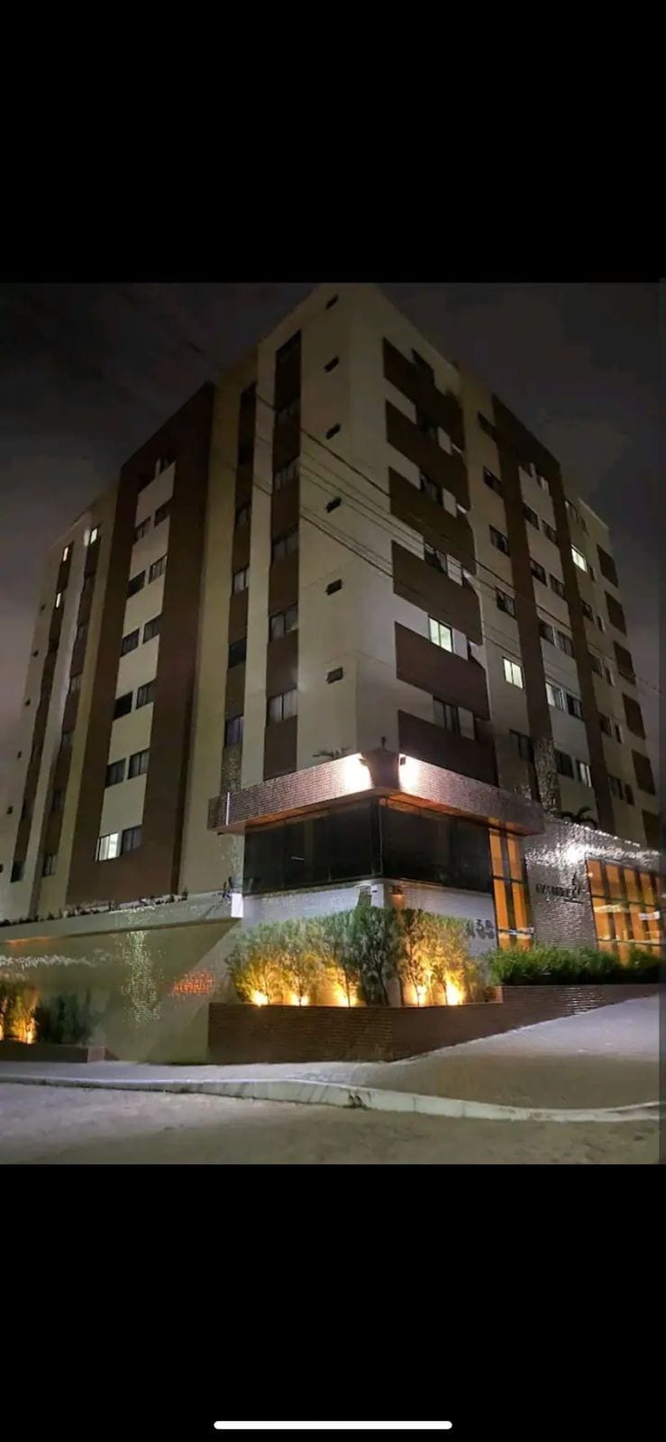 A modern multi-story building is illuminated at night, showcasing a combination of light and dark facades. Soft lighting highlights the entrance and landscaping, which features small plants and greenery along the base, creating an inviting appearance and providing warmth to the urban environment.