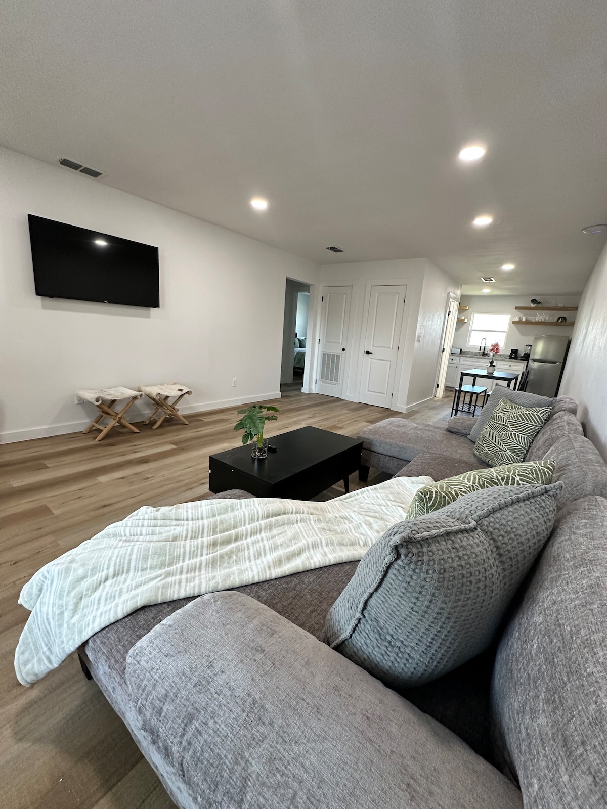 A welcoming living area features a spacious gray sectional sofa adorned with decorative pillows and a soft throw. A dark coffee table sits in front, while a wall-mounted TV provides entertainment options. Bright lighting enhances the light-colored walls, creating an inviting atmosphere.