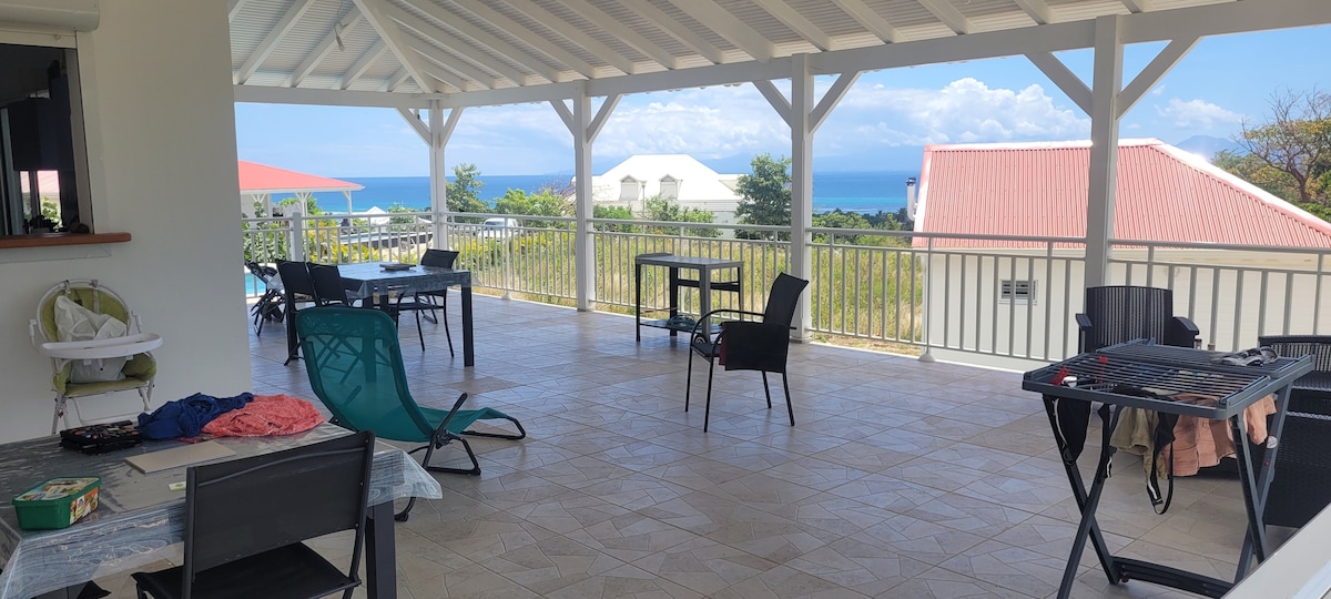 A spacious terrace offers a panoramic view of the sea, furnished with multiple tables and chairs. The open roof allows natural light to fill the area, while a few shaded spots provide comfort. Soft, colorful fabric drapes casually on one table, adding subtle detail.
