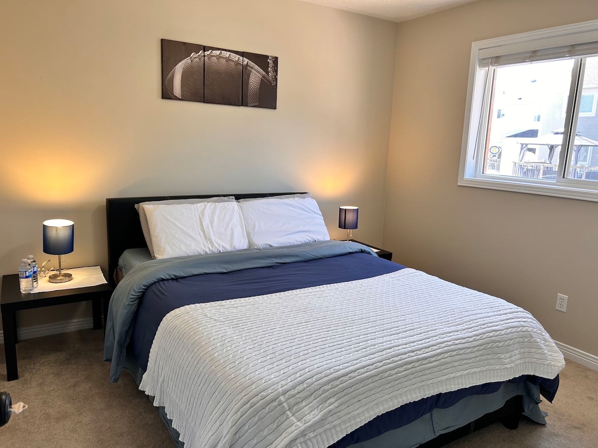 A bright bedroom features a spacious bed dressed in a white textured blanket, with blue linens at the foot. Two bedside tables with lamps provide soft lighting, and a window allows natural light to fill the room, framed by light-colored walls.