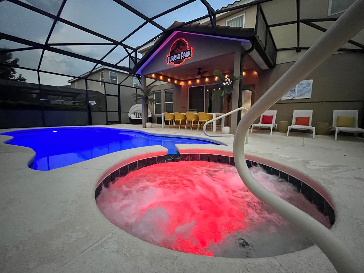 A heated spa is positioned next to a vibrant blue pool, both contained within a screened enclosure. Colorful lights illuminate the spa's water. Comfortable outdoor seating is arranged under a covered area, providing shaded relaxation near the pool.