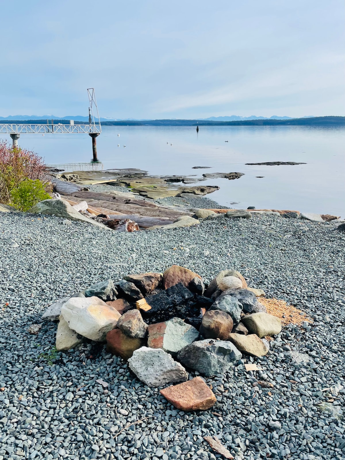 A fire pit composed of varied stones is situated on a rocky shoreline, with gentle waves lapping at the water's edge. A peaceful view of the calm sea and distant mountains is visible in the background, providing a serene setting for gatherings.