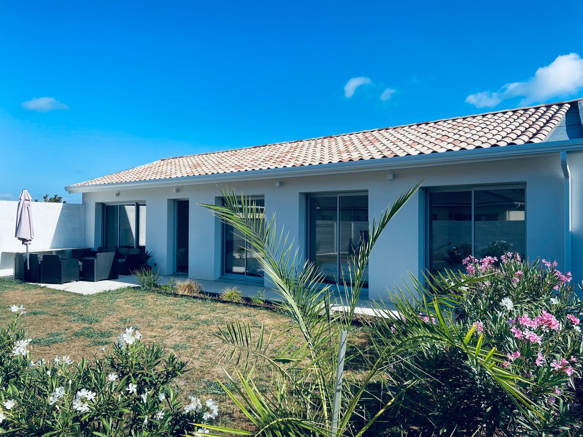 A modern house is viewed from the garden, featuring a light-colored exterior and a tiled roof. Large windows allow natural light into the interior. Lush green plants and flowering shrubs add color to the landscape, while a shaded seating area is visible on the patio.