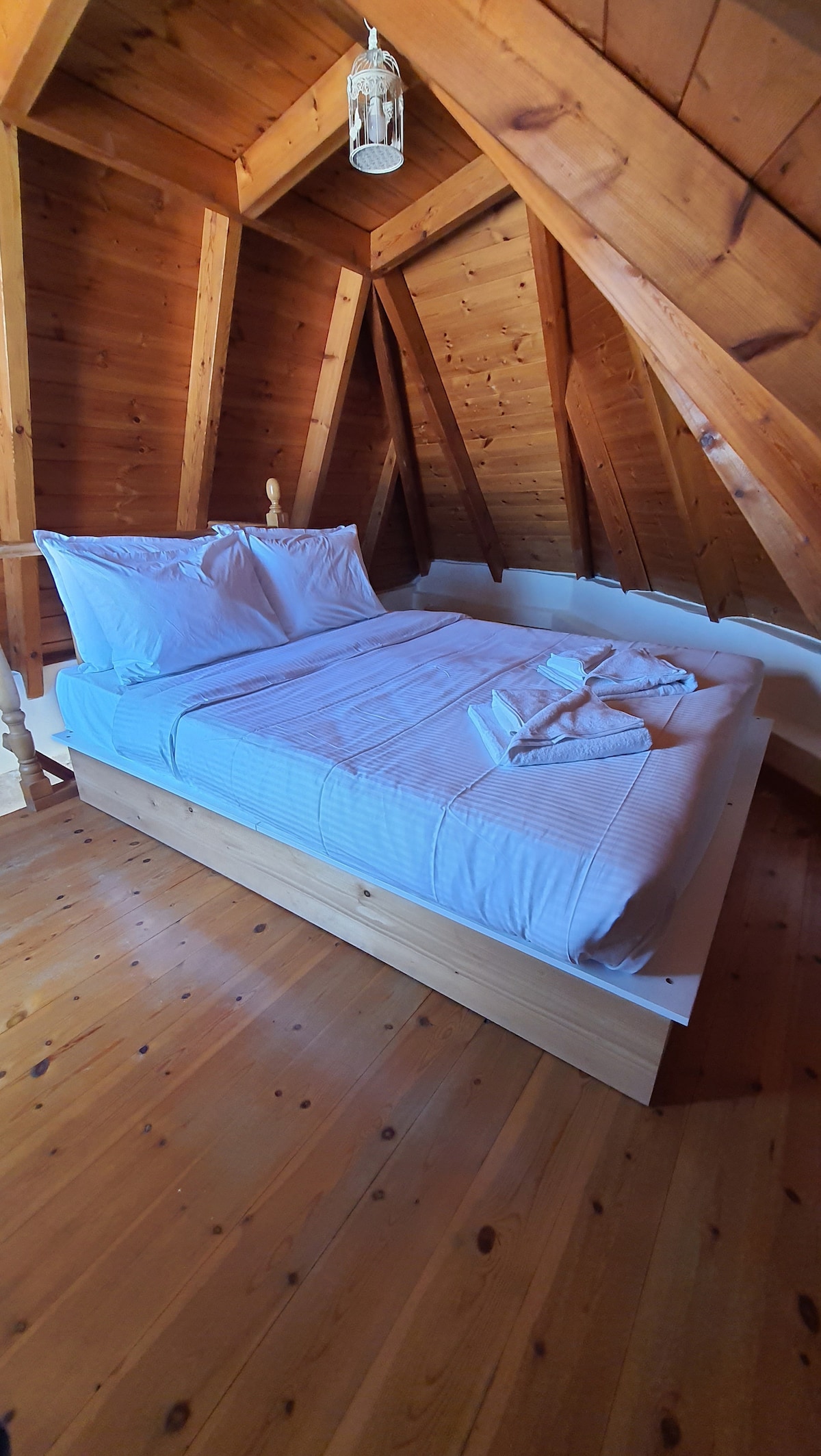 A cozy loft space features wood-paneled walls and an A-frame ceiling. A spacious bed is positioned centrally, adorned with crisp white linens. Natural light filters through a nearby window, highlighting the wooden flooring and creating a warm ambiance.