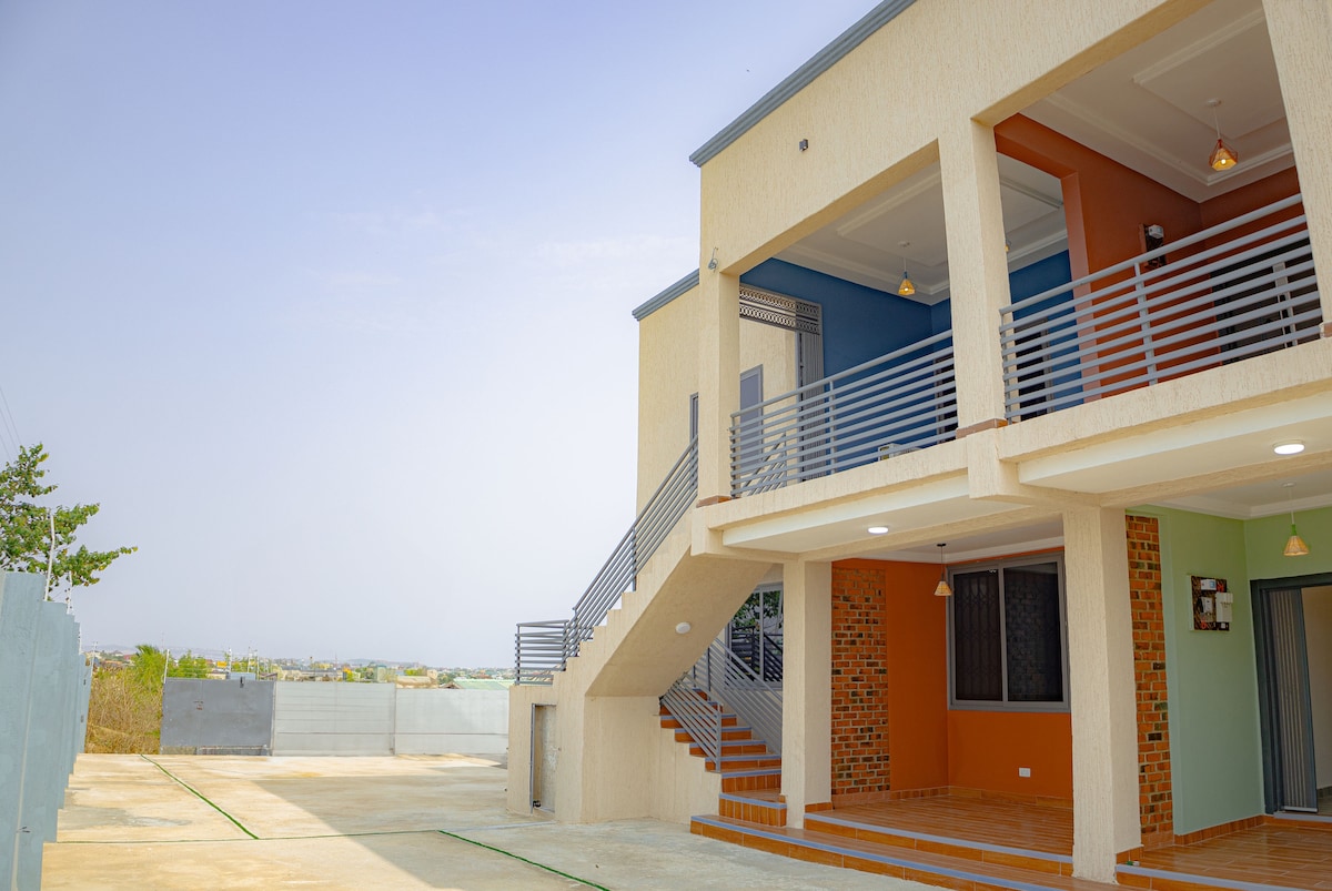The exterior of the two-story apartment is shown, featuring a mixture of light-colored façades and contrasting vibrant colors like orange and teal. A staircase leads to the upper floor, while ample outdoor space surrounds the building, offering a clear view of the serene environment.