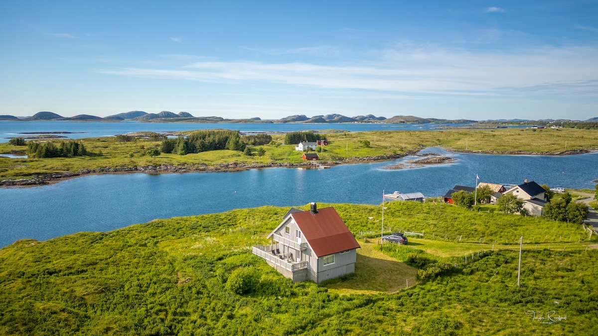 A charming house is situated on a lush green landscape near the water, surrounded by hills and scenic views. The structure features a red roof and a balcony, offering expansive views of the nearby bay and islands in the distance.