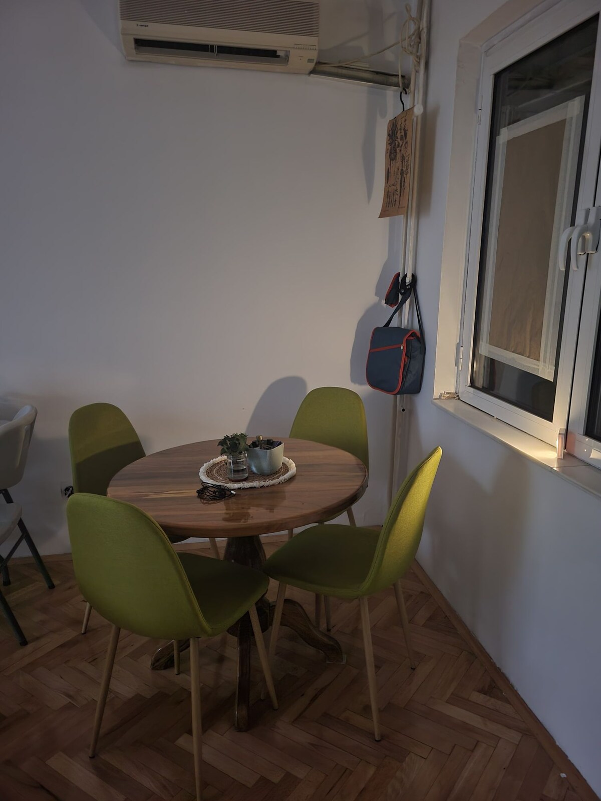 A round wooden table is accompanied by four green chairs, creating a cozy dining space. A small potted plant is positioned on the table, while natural light filters through a nearby window, illuminating the warm wooden floor.